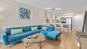 Una sala moderna con un sofá azul, una mesita de centro de vidrio y una cocina al fondo.