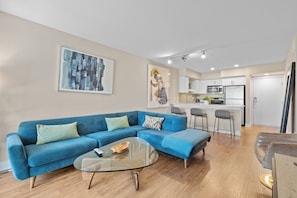A modern living room with a blue sofa, a glass coffee table, and a kitchen in the background. - Del Mar Direct access to the main channel Water View, Pool, jacuzzi and Gym (Marina del Rey)