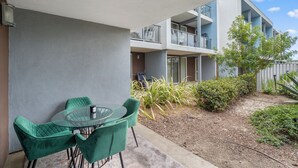 Un balcón con una mesa y sillas de vidrio, vista a un edificio con balcones y algo de vegetación.