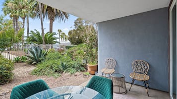 A patio with a glass table and two wicker chairs, surrounded by a garden with palm trees and various plants.
