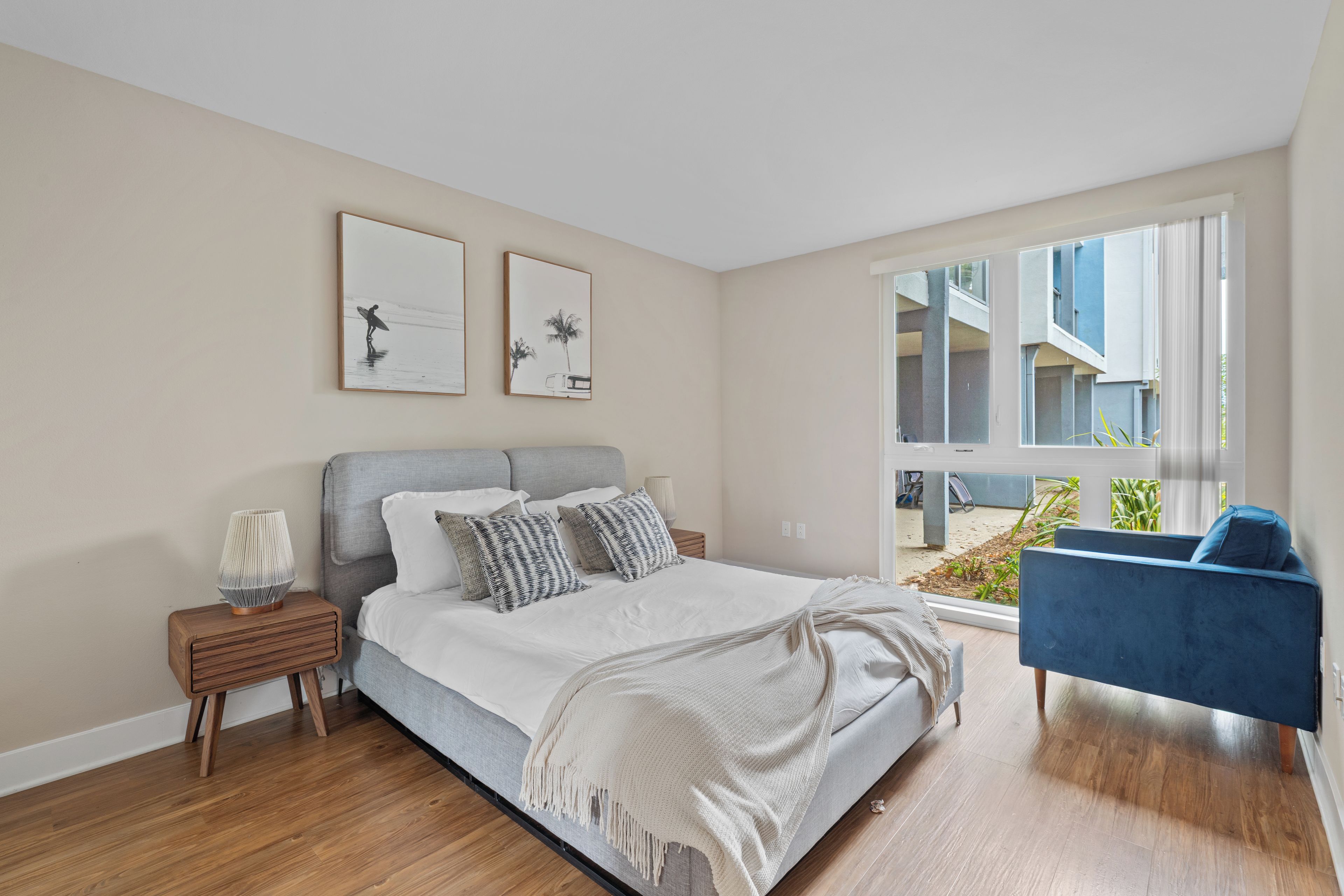 A bedroom with a bed, a nightstand, a lamp, a chair, and a large window with a view of buildings.