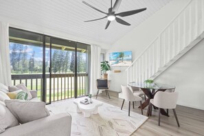 Ein modernes Wohnzimmer mit einer Couch, Couchtisch und Sesseln, einem Glastisch im Essbereich und einem Balkon mit Blick auf Bäume und Himmel.