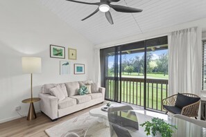 A modern living room with a sofa, coffee table, and a view of a green landscape through large windows. - ~NEW~ Exclusive Beach Loft ~Turtle Bay Golf Course (Kahuku)