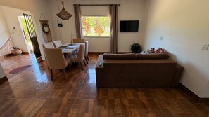 Una sala de estar con un sofá, una mesa de comedor con sillas, un televisor montado en la pared y un ventanal con cortinas.