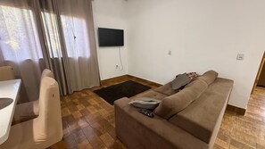 A living room with a sofa, a TV mounted on the wall, a dining table, and a window with curtains.