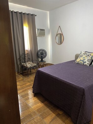 A bedroom with a bed, a chair, a small table, a fan, and a mirror on the wall.