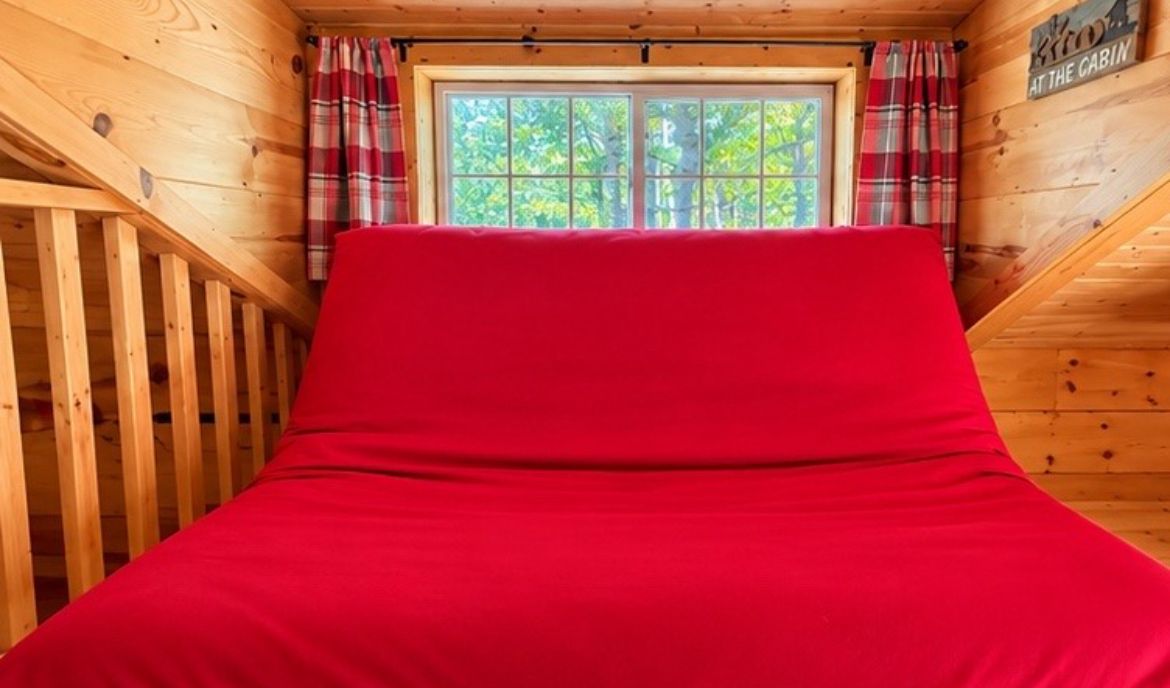 A cabin interior with a red bed, wooden walls, a window with curtains, and a sign reading 'At the Cabin'.