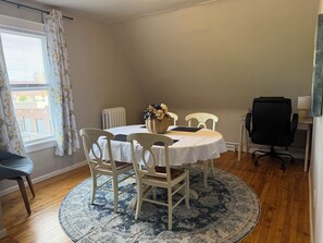 A dining area with a round table, white chairs, a vase with flowers, a radiator, and a window with curtains.
