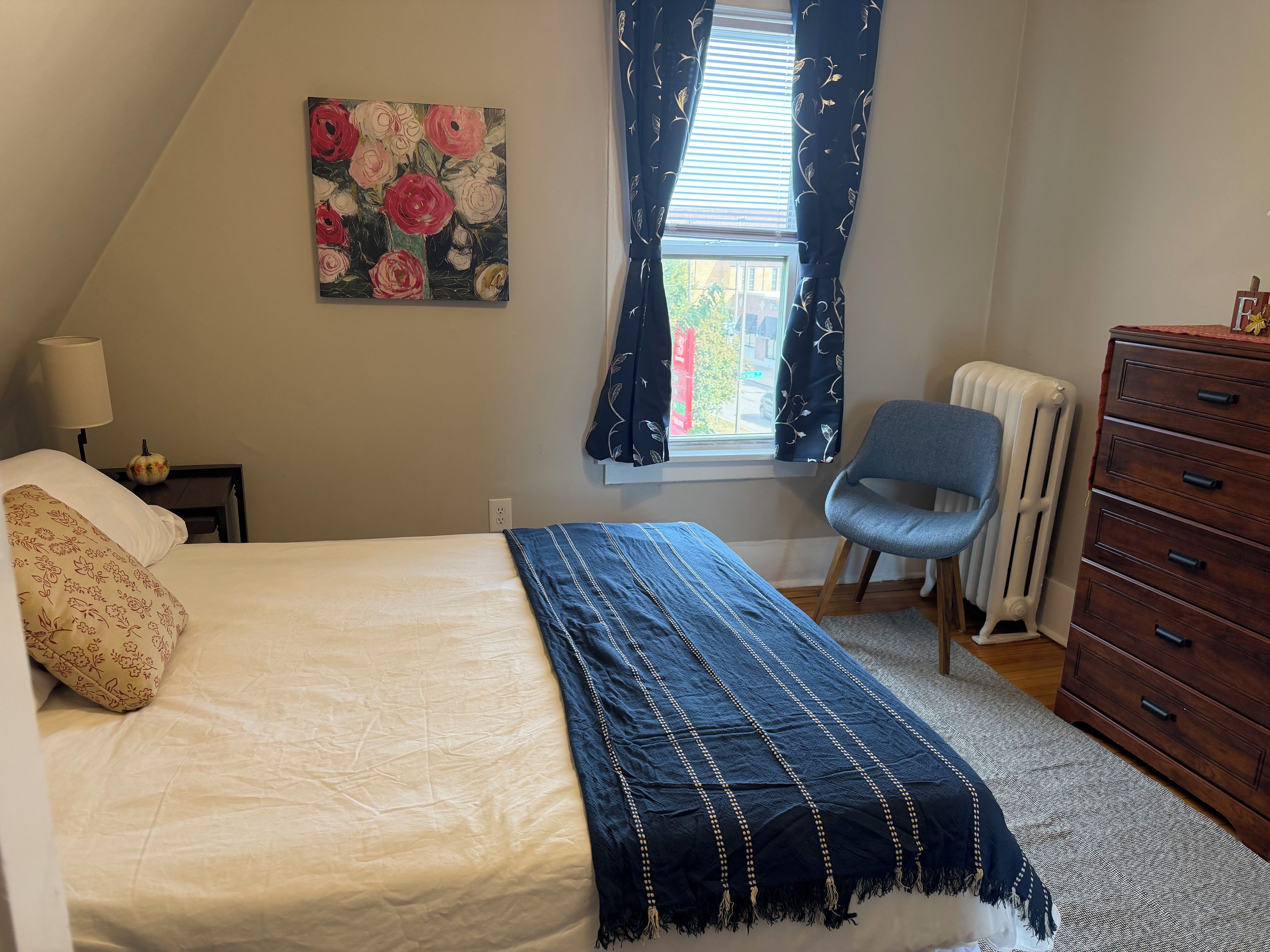 Een slaapkamer met een bed, een stoel, een dressoir, een radiator en een raam met gordijnen.