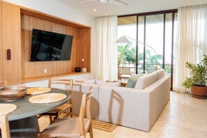 Ein modernes Wohnzimmer mit einem an einer Holzwand montierten Flachbildfernseher, einer Couch, einem Esstisch mit Stühlen und einem großen Fenster mit Vorhängen.