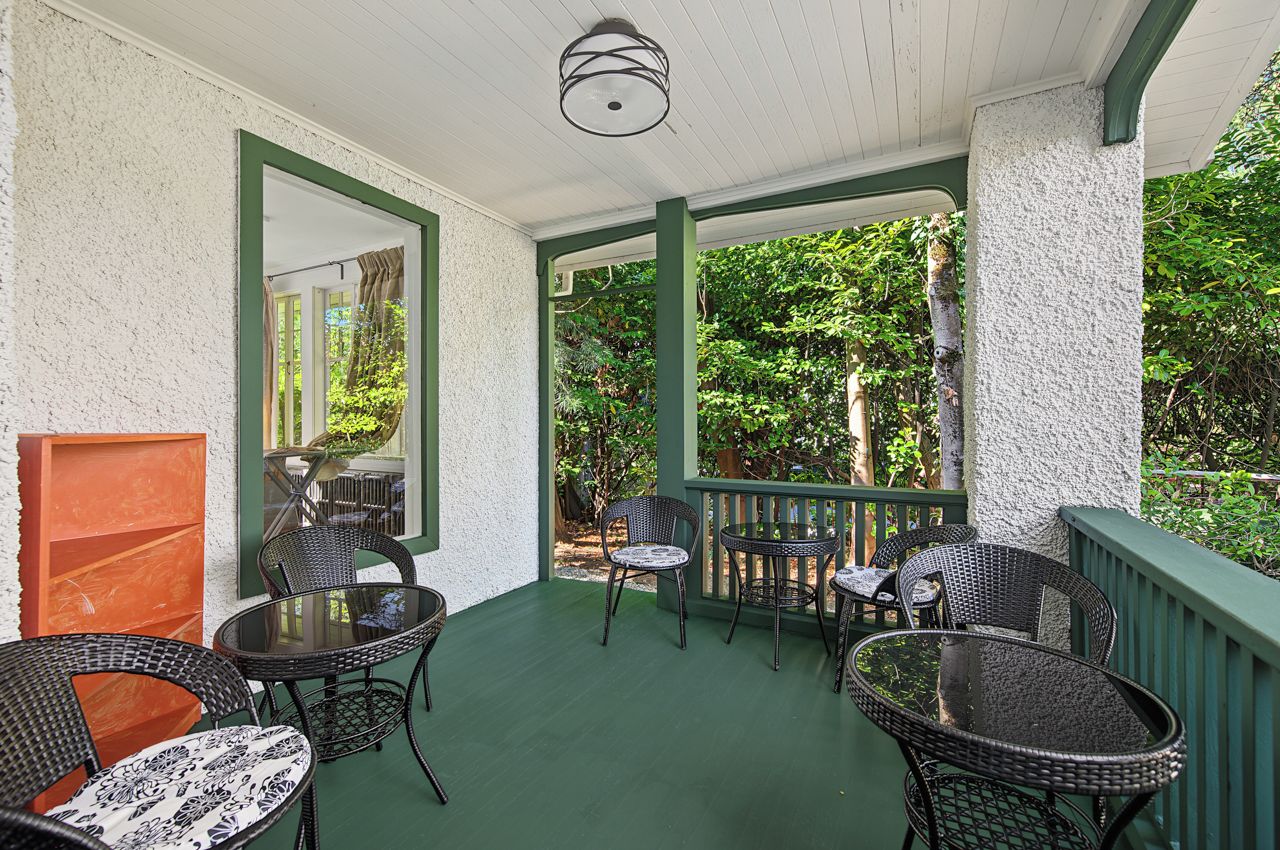 A covered outdoor area with wicker furniture, a glass table, and a view of greenery.
