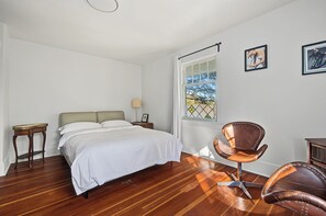 Un dormitorio con cama, una silla, mesita de noche, lámpara y ventana con vistas.