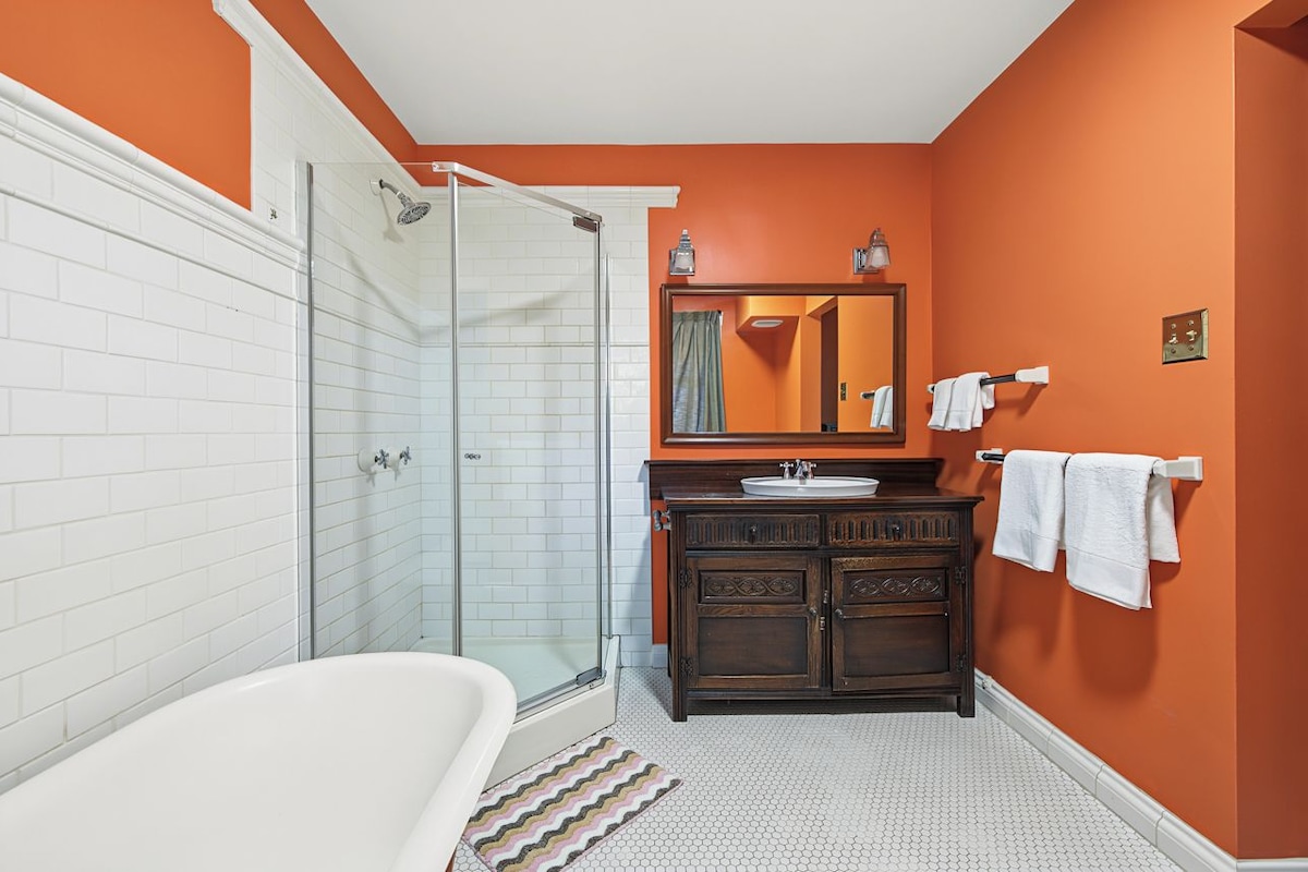 Een badkamer met een glazen douchecabine, een witte badkuip, een houten wastafelmeubel met een spiegel en oranje wanden.