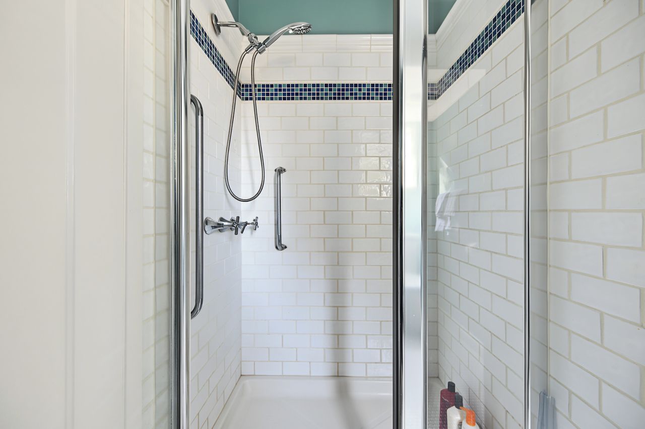 A shower with a glass door, white tiled walls, and a blue-tiled accent strip.
