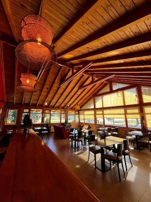 A spacious indoor area with wooden beams and large windows, featuring tables and chairs arranged for dining.