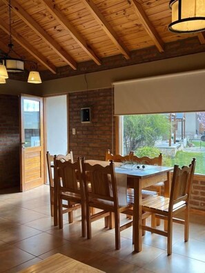A dining area with a wooden table and chairs, a brick wall, and a window with a view of the outdoors. - Hostería y Cabañas Land Park (Junín de los Andes)
