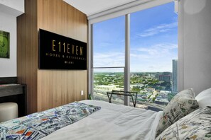 A hotel room with a large window offering a city view, a bed with a patterned bedspread, and a sign indicating the hotel name and location. - Deluxe E11EVEN Suite with Elite Amenities (Miami)