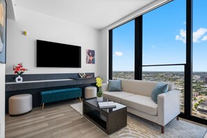 Ein modernes Wohnzimmer mit großem Fernseher, einer Couch, einem Couchtisch und Blick auf die Stadt durch große Fenster.