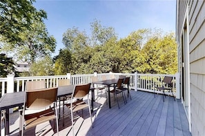 An outdoor deck with tables and chairs, surrounded by a white fence and trees. - Walk the beach from a spectacular 9-bed 6-bath Estate with sweeping ocean views. (Narragansett)