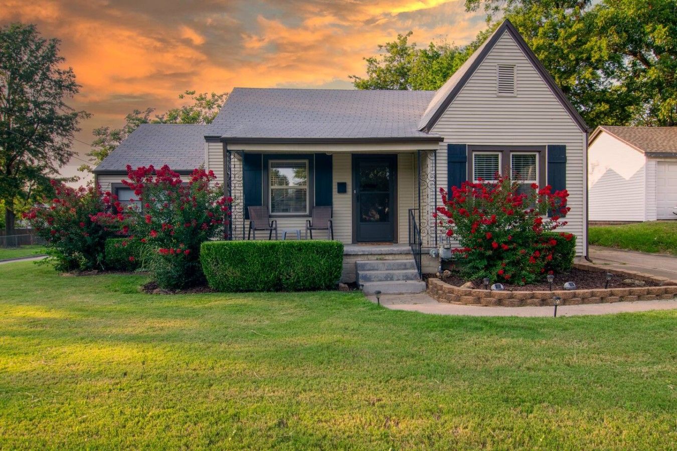 A single-story house with a front porch, a small garden with red flowers, and a clear sky.