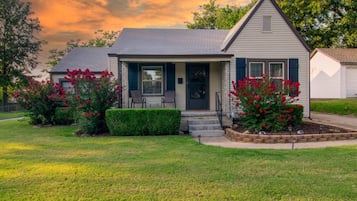 A single-story house with a front porch, a small garden with red flowers, and a clear sky.