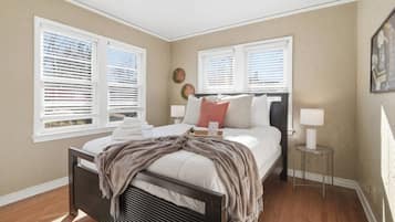 A bedroom with a bed, two bedside tables, a window with blinds, and a wall-mounted lamp.