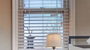 A bedroom with a bed, a bedside table with a lamp, and a window with blinds. - The Corner Charmer• Near TU/Expo/Downtown (Tulsa)