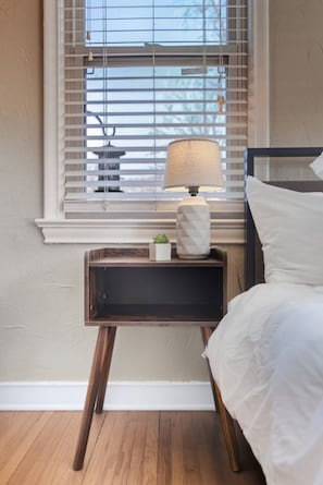 Een slaapkamer met een bed, een nachtkastje met een lamp en een raam met jaloezieën.