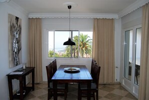 A dining area with a table, chairs, a pendant light, a window with curtains, and a painting on the wall. - Apartment in Nerja Near Burriana Beach (Nerja)