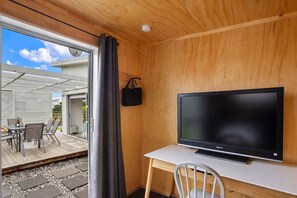 Quarto com teto de madeira, TV de tela plana em escrivaninha branca, cadeira e vista para um pátio externo com mesa e cadeiras.