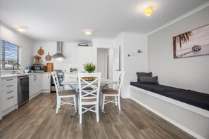 A modern kitchen with white cabinetry, a dining table with chairs, and a built-in oven. - Whanga on Foot - Brilliant Location (Whangamatā)