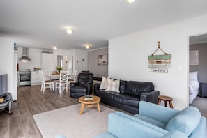Uma sala de estar moderna com um sofá de couro preto, um sofá azul e uma mesa de centro de madeira. Há uma área de jantar com cadeiras brancas e uma mesa, e uma cozinha ao fundo.