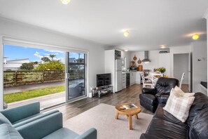 A modern living room with a sofa, coffee table, and a view of the backyard. - Whanga on Foot - Brilliant Location (Whangamatā)
