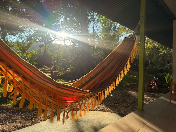 A hammock under a shaded area with a view of trees and a garden.