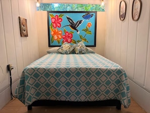 A bed with a patterned bedspread, a painting of birds and flowers on the wall, and a power outlet on the wall.