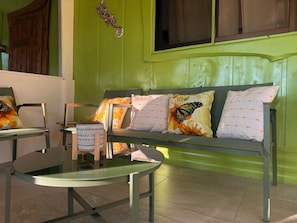 A room with a green wall, a sofa with butterfly-themed pillows, a glass table, and a wooden chair.
