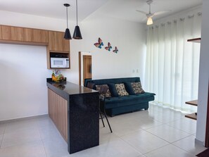 A modern kitchen with a black island, wooden cabinets, a microwave, and a fruit bowl. There is a teal sofa with patterned cushions and a ceiling fan. - Duplex Apartment 2 with 2 Suites 450m from the Beach in Santa Cruz Cabrália (Santa Cruz Cabrália)