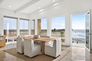 Ein geräumiges Zimmer mit großem Tisch und Stühlen, großen Fenstern mit Blick auf den Strand und einer Tür, die nach draußen führt.