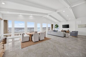 Een ruime woonkamer met een groot raam met uitzicht op de oceaan, een eetgedeelte met een houten tafel en een moderne keuken met een flatscreen-tv.