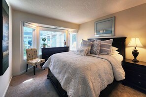Quarto com cama, cadeira, mesa de cabeceira, abajur e janela com vista.
