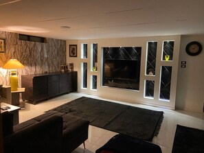 Ein modernes Wohnzimmer mit Fernseher, dunklen Sofas, einem Couchtisch, einer Lampe und einer Wand mit Marmorstruktur.