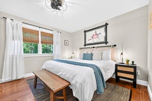 Ein Schlafzimmer mit einem Bett, Nachttischen, einer Bank, einem Deckenventilator, einem Fenster mit Vorhängen und einem gerahmten Bild an der Wand.