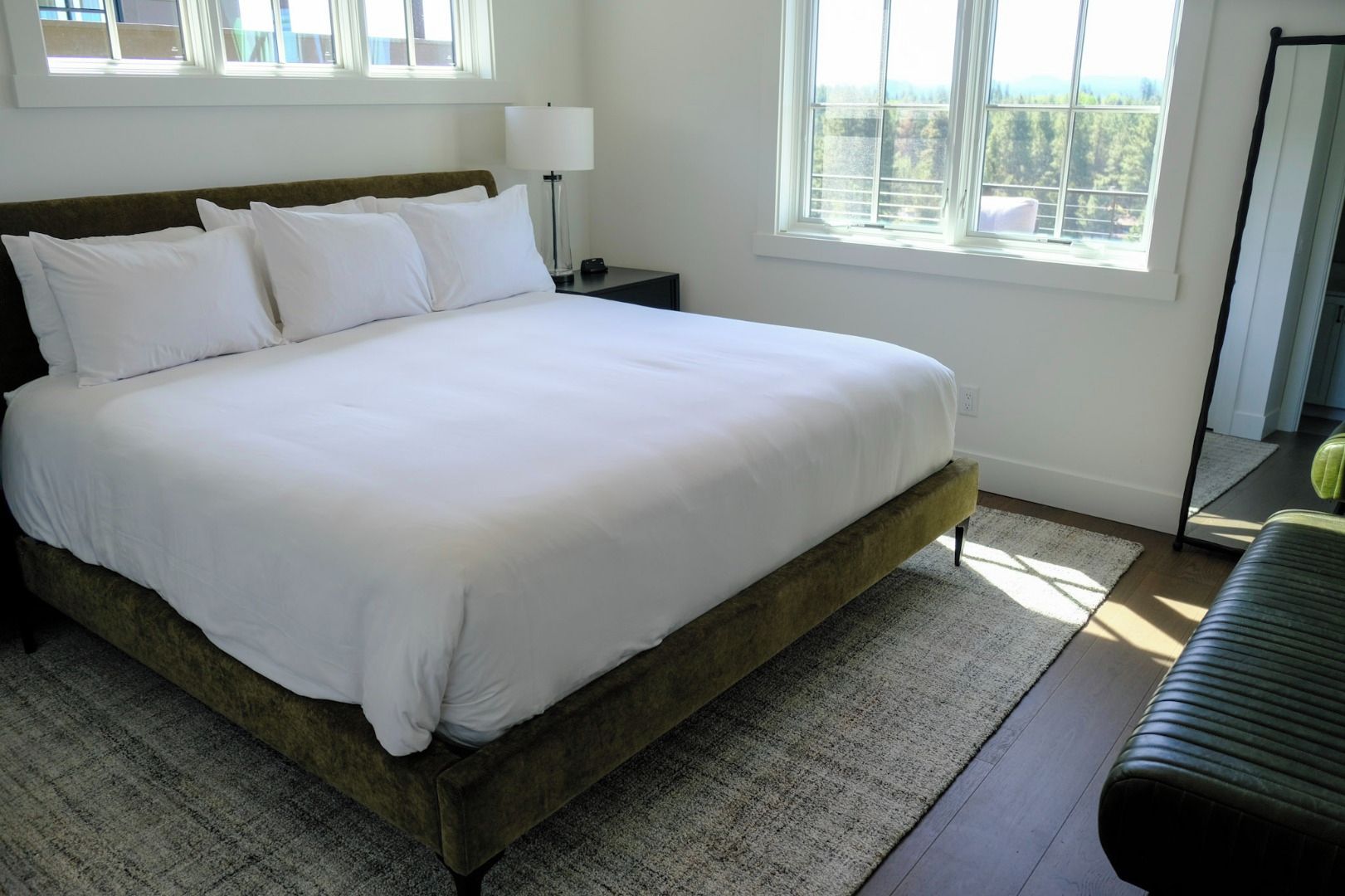 A bed with white bedding, a bedside lamp, and a view of trees through a window.