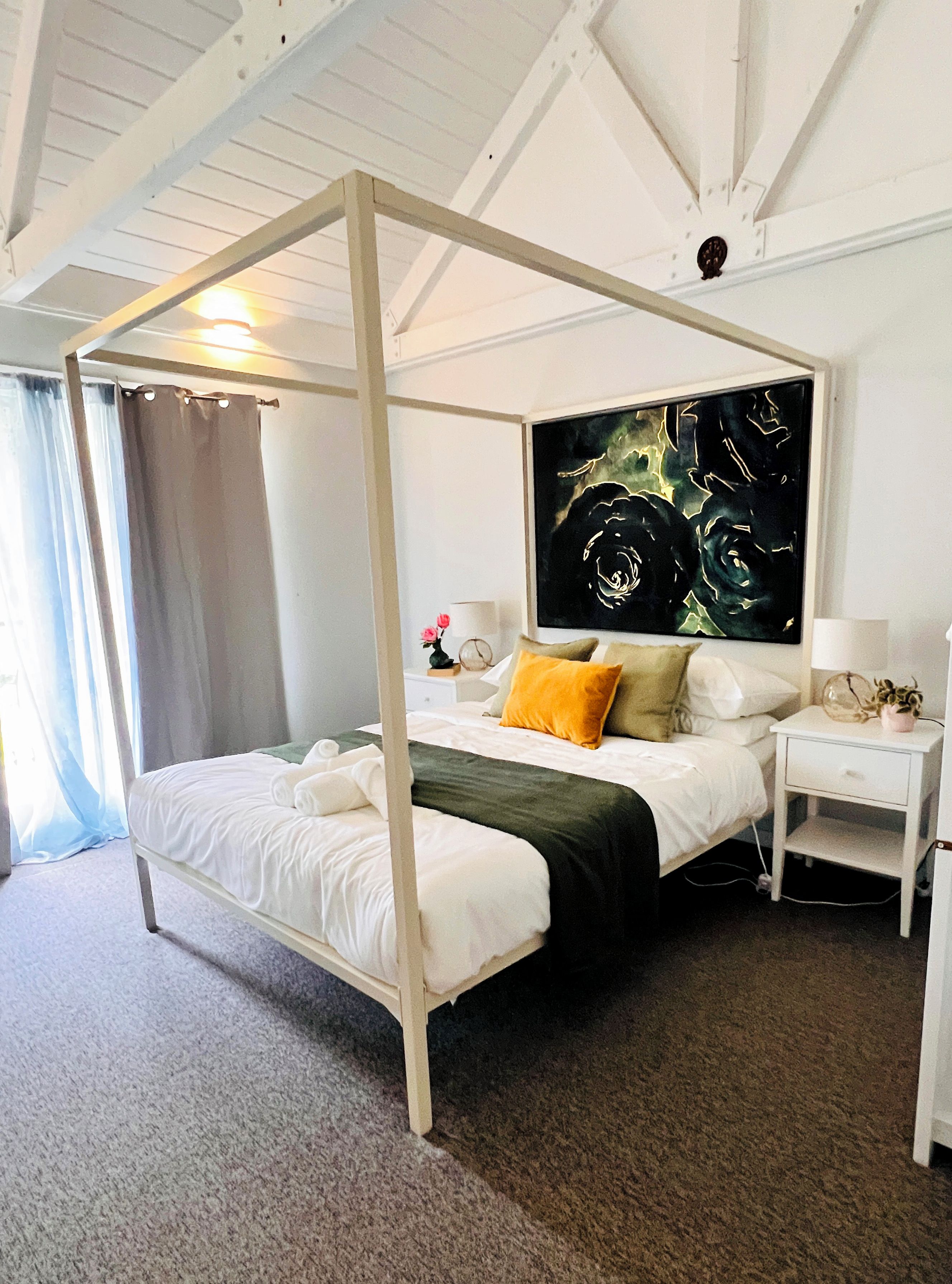 A four-poster bed with a dark green bedspread, white bedding, and a single yellow pillow. A large abstract painting is on the wall above the bed.