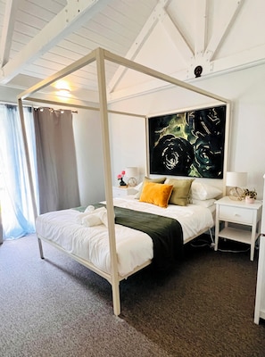 A four-poster bed with a dark green bedspread, white bedding, and a single yellow pillow. A large abstract painting is on the wall above the bed. - McLaren Vale Vineyard Loft | Full Kitchen + Bath (Blewitt Springs)