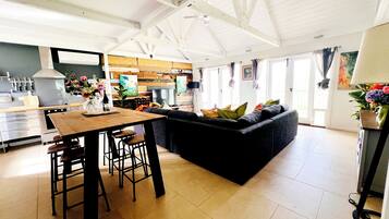 A spacious living area with a dark-colored sofa, a wooden dining table with stools, and a kitchen in the background.