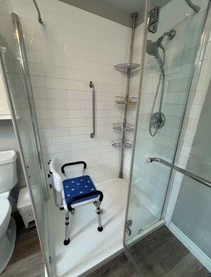 A shower area with a glass door, a white tiled wall, a blue wheelchair-accessible bench, and a handheld showerhead.