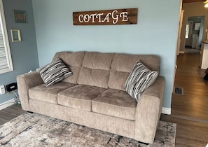 A beige sofa with striped pillows, a rustic 'COTTAGE' sign, and a patterned rug in a room with light blue walls and wooden flooring.