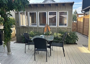 A wooden deck with a glass table and chairs, surrounded by plants and a wooden fence.