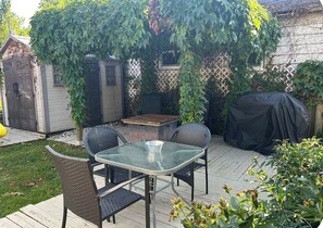 An outdoor patio with a glass table and wicker chairs, surrounded by greenery and a wooden deck.